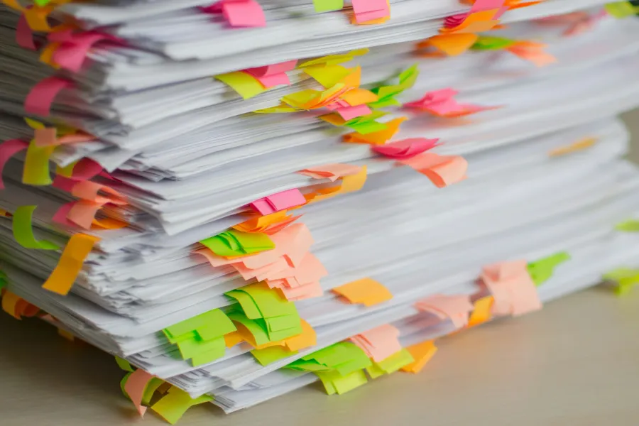 Ein hoher Stapel Papier auf einem hellen Schreibtisch, aus dem an jeder zweiten Lage bunte Haftstreifen in Pink, Orange, Gelb und Grün hervorstehen