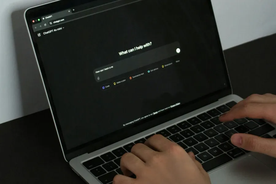 Zwei Hände tippen auf einem Laptop, auf dessen Bildschirm die Startseite von ChatGPT mit der Frage „What can I help with?“ geöffnet ist