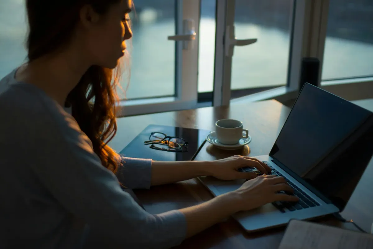 Eine Frau tippt am Abend am Laptop, daneben ihre Brille und eine Tasse