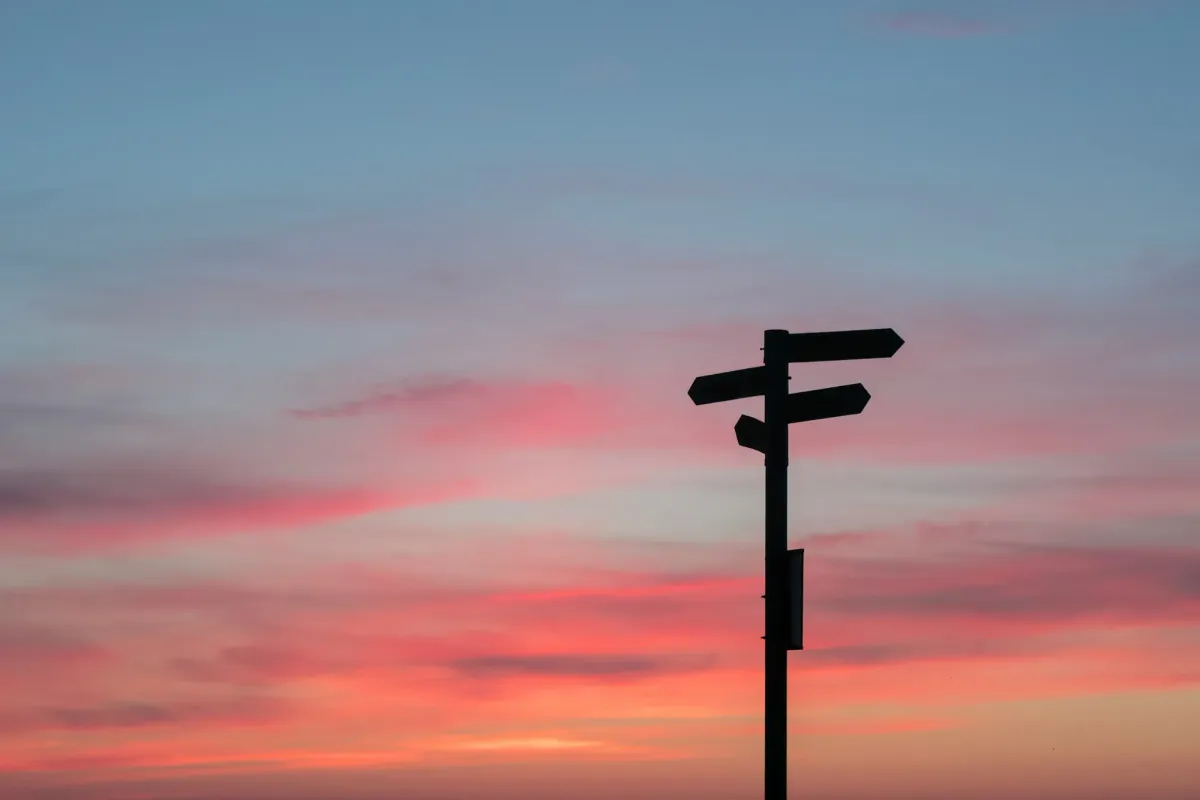Ein Wegweiser als Silhouette gegen einen rot-blauen Abendhimmel, dessen drei Schilder in drei verschiedene Richtungen zeigen