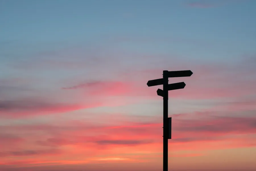 Ein Wegweiser als Silhouette gegen einen rot-blauen Abendhimmel, dessen drei Schilder in drei verschiedene Richtungen zeigen