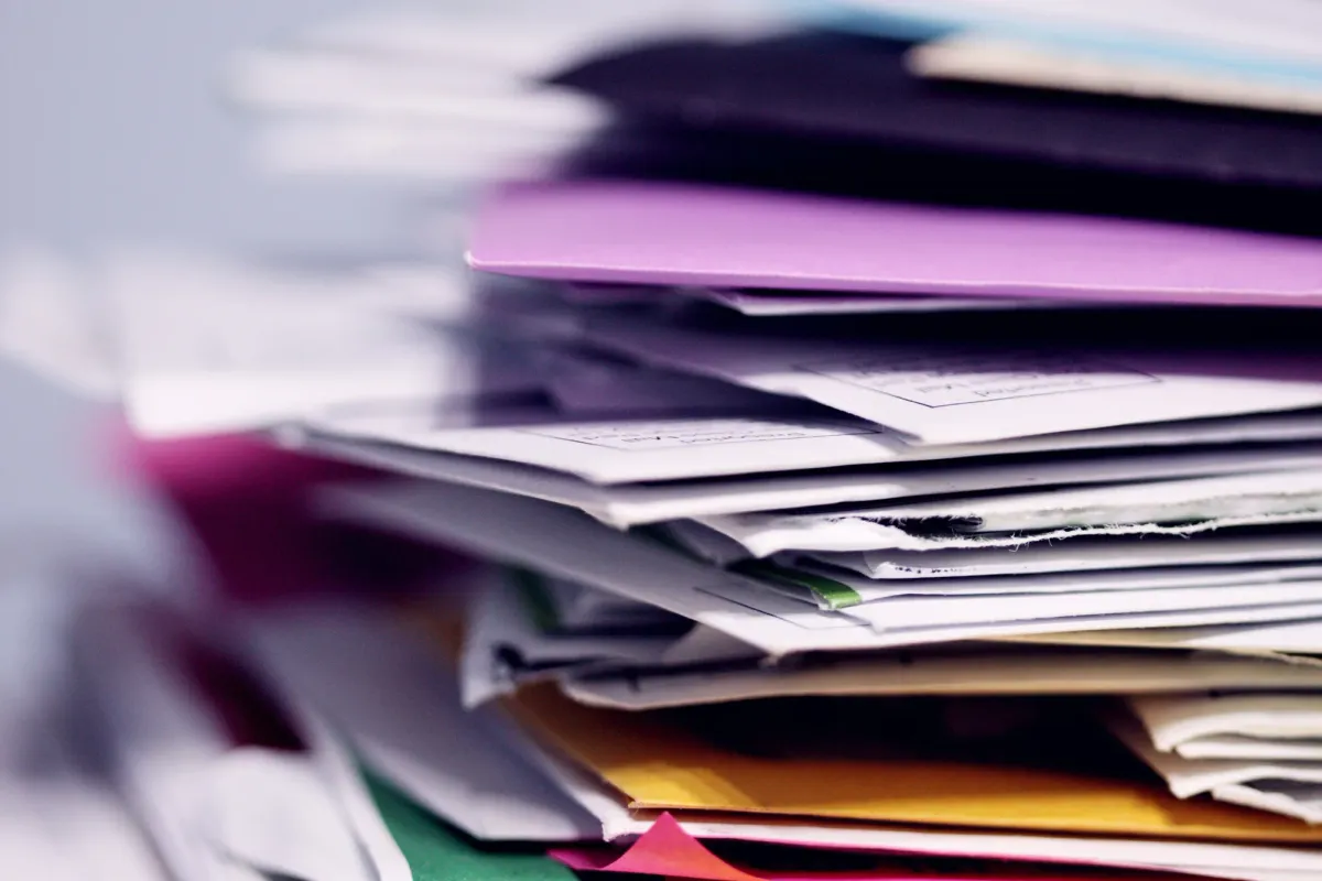 Ein unordentlicher Stapel aus Papieren, Umschlägen und farbigen Mappen in Lila, Gelb, Pink und Grün, von der Seite fotografiert, nach links in die Unschärfe auslaufend