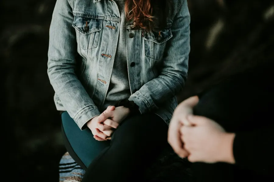 Zwei Menschen sitzen einander in einem abgedunkelten Raum gegenüber; im Licht eine Person in heller Jeansjacke mit ineinandergelegten Händen, im unscharfen Vordergrund die Hände des Gegenübers