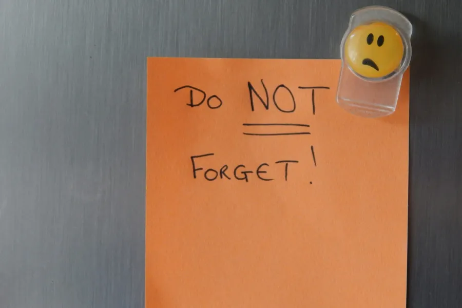 Ein orangefarbener Notizzettel an einer Kühlschranktür aus gebürstetem Metall, darauf handschriftlich „Do NOT forget!“, gehalten von einem gelben Magneten mit traurigem Gesicht